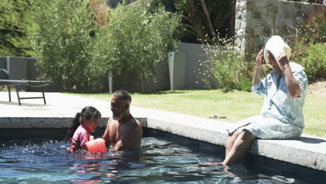 Biracial-man-and-child-enjoy-pool-time,-woman-in-a-hat-sits-beside-them