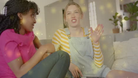 Animation-of-yellow-light-spots-over-two-happy-diverse-teenage-girls-talking-at-home