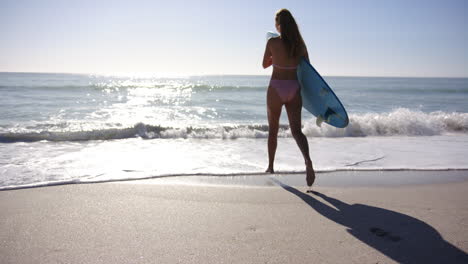 Young-Caucasian-woman-runs-towards-the-ocean-with-a-surfboard-under-her-arm,-with-copy-space
