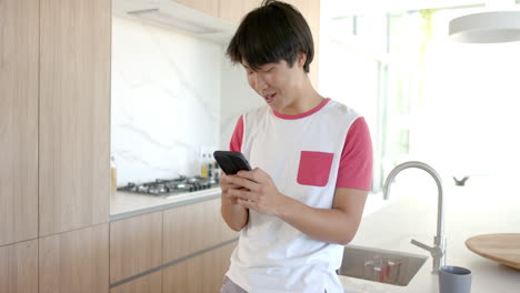 Un-Adolescente-Asiático-Sonríe-A-Su-Teléfono-En-Una-Cocina-Moderna.