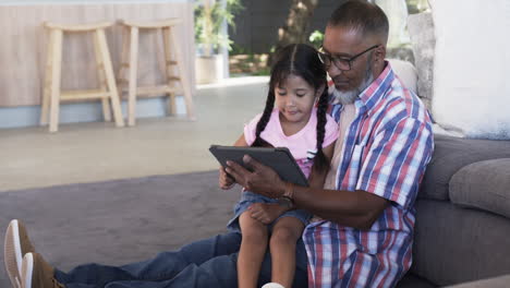 Biracial-grandfather-and-granddaughter-are-engrossed-in-a-tablet-while-sitting-on-a-couch