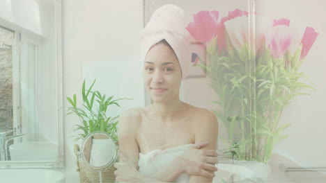 Animation-of-pink-flowers-in-vase-over-portrait-of-happy-biracial-woman-wearing-towel-in-bathroom