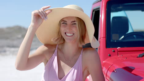 Young-Caucasian-woman-smiles-holding-her-hat-at-the-beach-on-a-road-trip