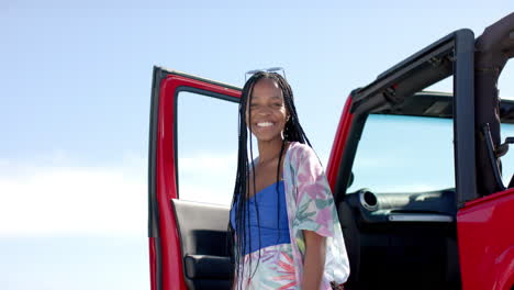 Young-African-American-woman-stands-by-a-red-car-outdoors-on-a-road-trip