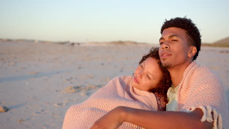Biracial-couple-embraces-wrapped-in-a-blanket-on-the-beach-at-sunset-with-copy-space