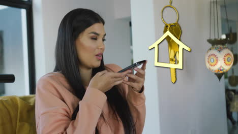 Animation-of-gold-house-key-and-key-fob-over-happy-biracial-woman-using-smartphone-at-home