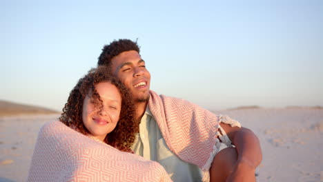Biracial-couple-wrapped-in-a-blanket,-enjoying-a-beach-at-sunset