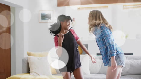 Animation-of-light-spots-over-two-happy-diverse-teenage-girls-dancing-at-home