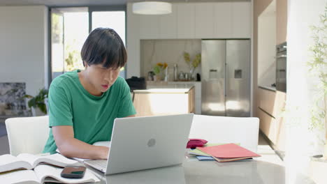 Adolescente-Asiático-Estudiando-En-Una-Computadora-Portátil-En-Una-Mesa-De-Cocina-En-Casa-Con-Espacio-Para-Copiar