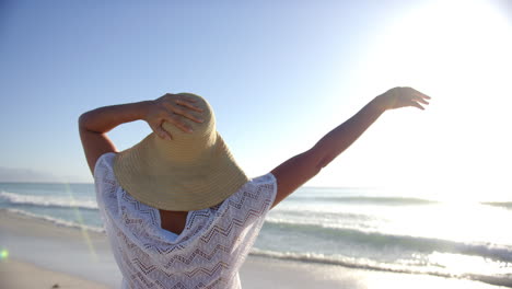 A-young-biracial-woman-enjoys-the-beach,-her-arms-outstretched-and-a-wide-brimmed-hat-on-her-head