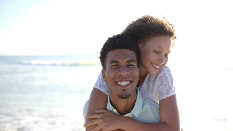 Biracial-couple-embraces-on-a-sunny-beach,-with-the-woman's-arms-wrapped-around-the-man's-shoulders