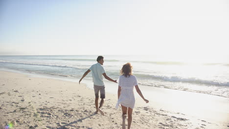 Biracial-couple-runs-along-a-sunlit-beach-with-copy-space,-their-backs-to-the-camera