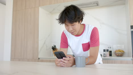 Asiatischer-Teenager-Lächelt,-Während-Er-Zu-Hause-In-Der-Küche-Sein-Smartphone-Benutzt