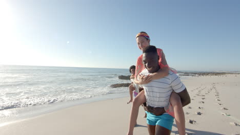 Young-African-American-man-gives-a-piggyback-ride-to-a-young-Caucasian-woman-on-the-beach