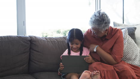 Biracial-girl-with-a-tablet-sits-beside-her-grandmother,-sharing-a-moment-of-joy-with-copy-space