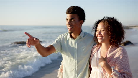 Biracial-couple-enjoys-a-sunny-beach-day,-the-man-pointing-towards-the-sea