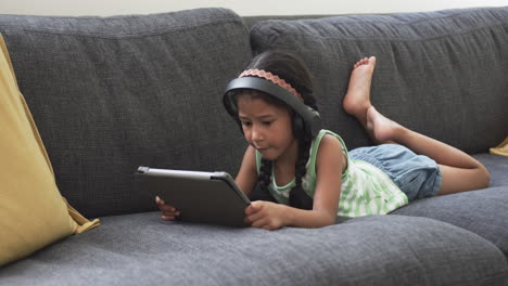 Biracial-girl-with-headphones-uses-a-tablet-while-lying-on-a-gray-couch