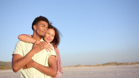 Biracial-couple-embraces-warmly-on-a-sunlit-beach,-eyes-closed-in-contentment,-with-copy-space