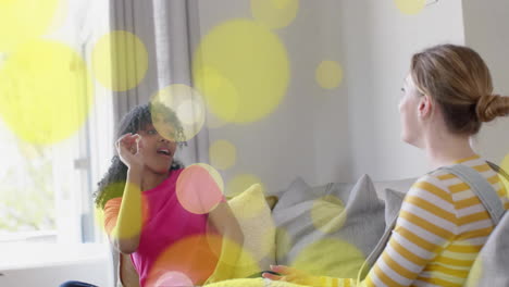 Animation-Von-Gelben-Lichtpunkten-über-Zwei-Unterschiedlichen-Teenager-Mädchen,-Die-Lachen-Und-Mit-Popcorn-Spielen