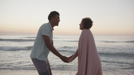 Biracial-couple-holds-hands-on-the-beach-at-sunset,-with-waves-in-the-background