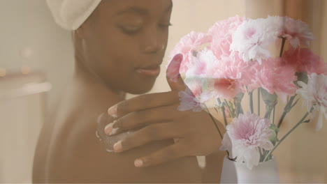 Animation-of-pink-flowers-in-vase-over-african-american-woman-moisturising-shoulder-in-bathroom