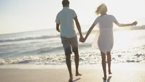 Biracial-couple-holding-hands,-walking-on-a-sunlit-beach
