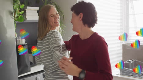 Animation-of-pride-rainbow-hearts-over-happy-caucasian-lesbian-couple-dancing-in-kitchen