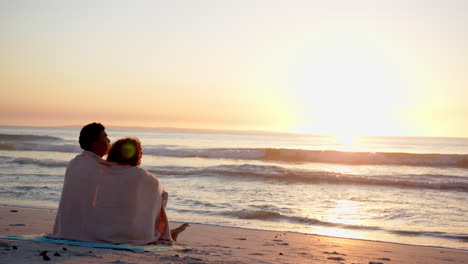 Biracial-couple-wrapped-in-a-blanket,-watching-a-sunset-at-the-beach-with-copy-space
