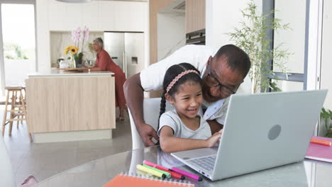 Biracial-grandfather-helps-a-young-biracial-granddaughter-with-a-laptop-in-a-modern-kitchen