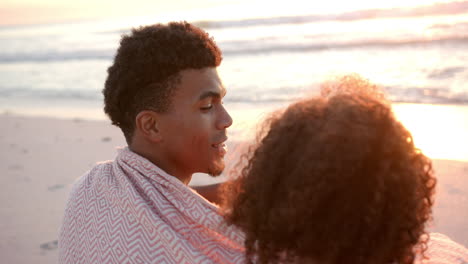 Biracial-couple-enjoys-a-serene-beach-sunset,-wrapped-in-a-cozy-blanket