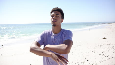 Young-biracial-man-stands-with-arms-crossed-on-a-sunny-beach