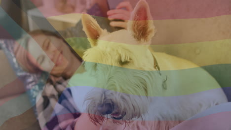 Animation-of-pride-rainbow-flag-over-happy-caucasian-woman-relaxing-with-smartphone-and-pet-dog