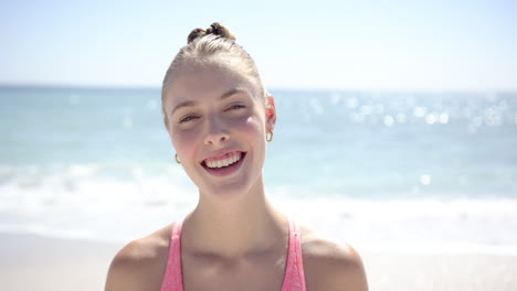 A-young-Caucasian-woman-with-blonde-hair,-wearing-a-casual-pink-top,-smiles-brightly-at-the-beach