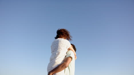 Biracial-couple-embraces-under-a-clear-blue-sky,-woman's-curly-hair-blowing-in-the-wind