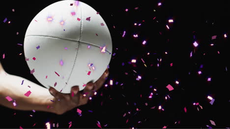 Animation-of-confetti-over-white-rugby-ball-held-by-caucasian-man's-hand-on-black-background