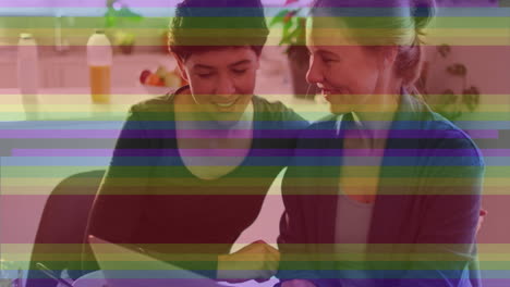 Animation-of-pride-rainbow-stripes-over-happy-caucasian-lesbian-couple-using-laptop-at-home
