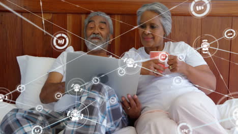 Animation-of-network-of-connections-with-icons-over-senior-biracial-couple-using-laptop-in-bed