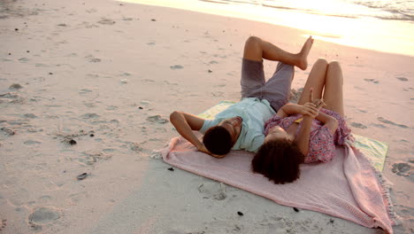 Biracial-couple-relaxes-on-a-beach-towel-at-sunset-with-copy-space