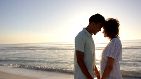 Biracial-couple-stands-close-on-a-beach-at-sunset,-foreheads-touching-affectionately,-with-copy-spac