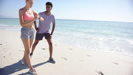 Young-Caucasian-woman-checks-her-smartwatch-beside-a-young-biracial-man-on-a-sunny-beach-with-copy-s