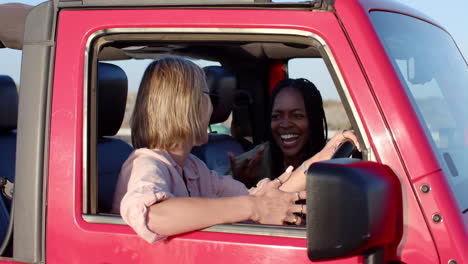 Young-African-American-woman-and-young-Caucasian-woman-share-a-laugh-in-a-car-on-a-road-trip,-with-c