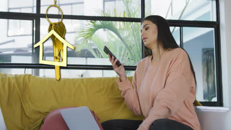 Animation-of-gold-house-key-and-key-fob-over-happy-biracial-woman-using-smartphone-at-home