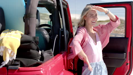 Young-Caucasian-woman-shields-her-eyes-from-the-sun-beside-a-red-car-on-a-road-trip