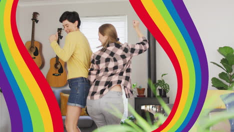 Animation-of-pride-rainbow-bands-over-happy-caucasian-lesbian-couple-dancing-at-home