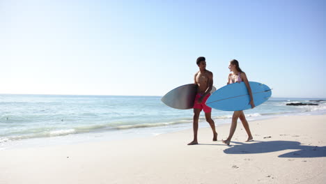 Biracial-man-and-Caucasian-woman-carry-surfboards-on-a-sunny-beach-with-copy-space