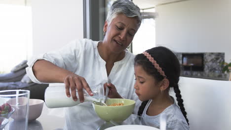 Biracial-grandmother-pours-milk-into-a-bowl-for-a-young-biracial-granddaughter-at-a-kitchen-table