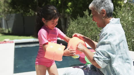 Biracial-girl-in-a-pink-swimsuit-pours-water-from-a-jug,-assisted-by-her-grandmother
