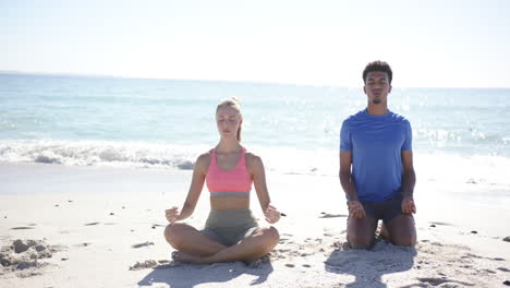 Young-Caucasian-woman-and-biracial-man-meditate-on-a-sunny-beach