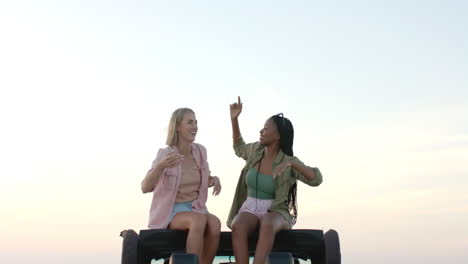 Young-African-American-woman-and-young-Caucasian-woman-sit-atop-a-vehicle-outdoors-on-a-road-trip