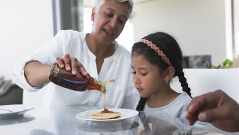 Biracial-grandmother-pours-syrup-on-a-pancake-for-a-young-biracial-granddaughter-at-a-kitchen-table